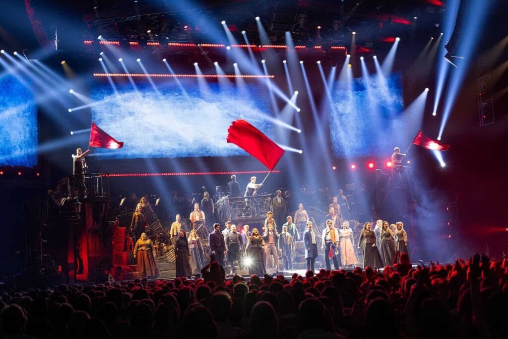 Podium vol met acteurs van Les Misérables The Arena Spectacular, waarbij er met een grote rode vlag en twee kleinere rode vlaggen wordt gezwaaid. Er zijn veel blauw-witte lichtbundels te zien, waarvan het grootste deel van achter het podium omlaag schijnt naar de voorkant van het podium, en er enkele naar boven en naar links of rechts schijnen. Voor het podium zie je een aantal rijen publiek.